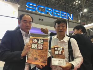 拡大版『絶景本棚』を持つメディアテクノロジージャパンの山岡さん（左）と、本の雑誌社の杉江さん。